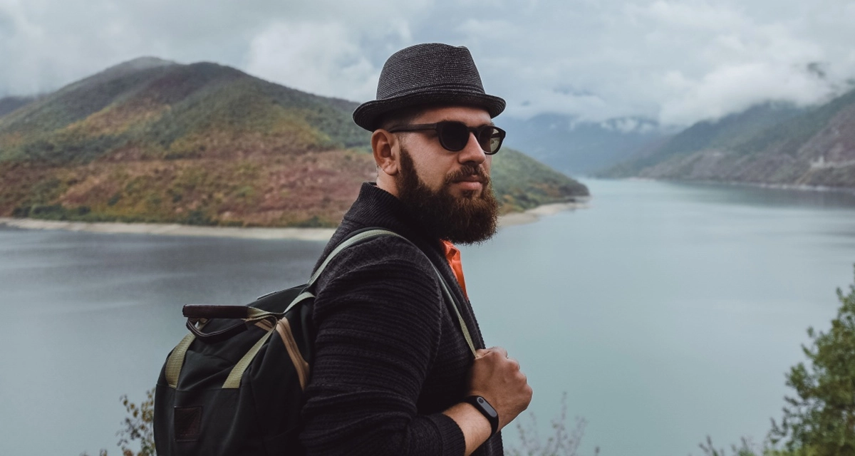 Bearded man wearing hat and sunglasses with backpack overlooking a lake and mountains on a cloudy day.
